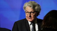 Thierry Breton listens with a serious expression, wearing a suit and glasses, in front of a blue background.