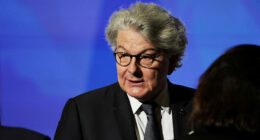 Thierry Breton listens with a serious expression, wearing a suit and glasses, in front of a blue background.