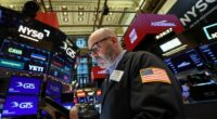 A trader in a jacket with a U.S. flag patch works on the NYSE floor, surrounded by digital stock ticker screens.