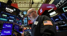 A trader in a jacket with a U.S. flag patch works on the NYSE floor, surrounded by digital stock ticker screens.