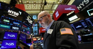 A trader in a jacket with a U.S. flag patch works on the NYSE floor, surrounded by digital stock ticker screens.