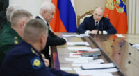 Vladimir Putin sits at the head of a long table during a meeting, with officials and documents in front of him. Russian flags are visible behind him.