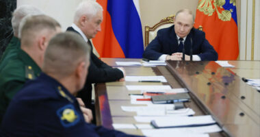 Vladimir Putin sits at the head of a long table during a meeting, with officials and documents in front of him. Russian flags are visible behind him.