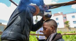 Barber and viral content creator Safari Martins, demonstrates one of his inventive shaving methods using an iron box while grooming Ian Njenga in Kiambu, Kenya, Wednesday, Nov. 26, 2025. (AP Photo/Andrew Kasuku)