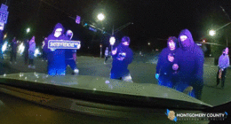 A mob blocks a police cruiser at a street takeover as smoke bombs explode in the background.