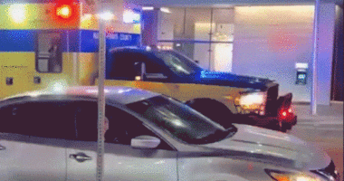 A Waymo autonomous car pauses near an emergency vehicle with flashing lights outside a downtown Austin garage at night.