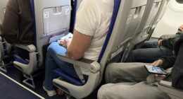 Passengers seated inside an airplane cabin with visible seat belts, legroom, and personal belongings during a commercial flight