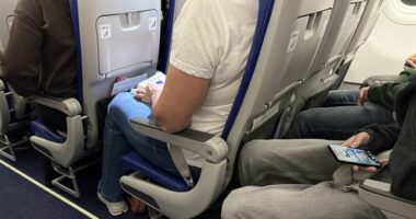 Passengers seated inside an airplane cabin with visible seat belts, legroom, and personal belongings during a commercial flight