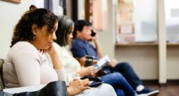 People sit in doctor's waiting room.  One woman looks a medical brochure and a man uses his smart phone to make a call.